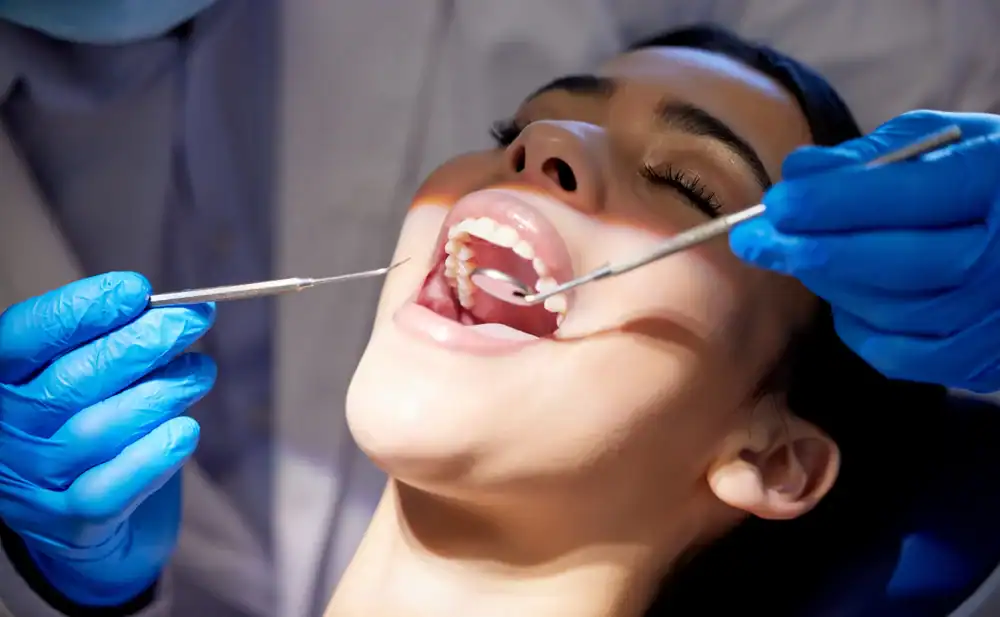A person reclines with their mouth open while a dentist, wearing blue gloves, examines their teeth using dental tools.
