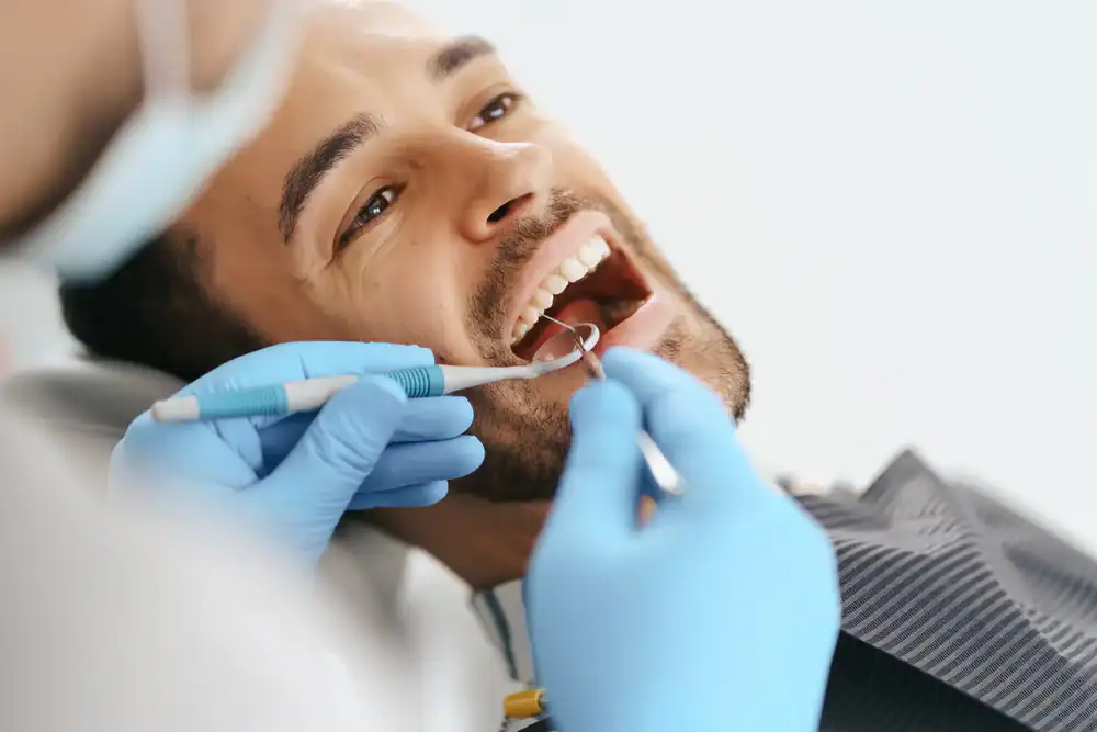 A man lies back in a dental chair with his mouth open while a dentist wearing blue gloves examines his teeth using dental instruments, showcasing a routine dentist visit.