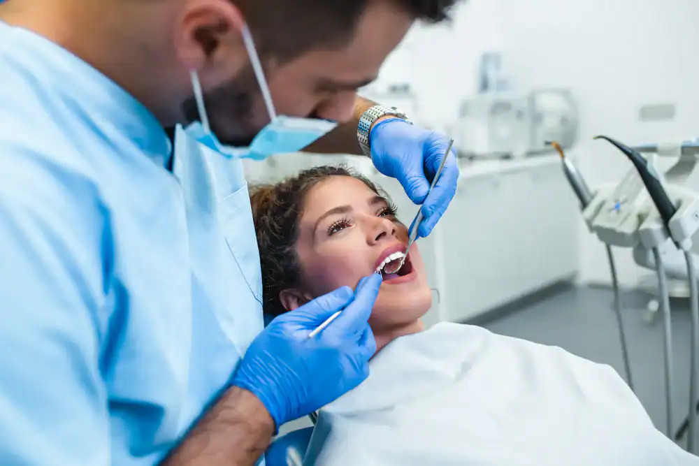 A dentist wearing a mask and gloves examines a woman’s teeth with dental tools as she sits in a dental chair, covered with a protective bib, in a modern dental clinic. The attentive dentist ensures her comfort throughout the checkup.