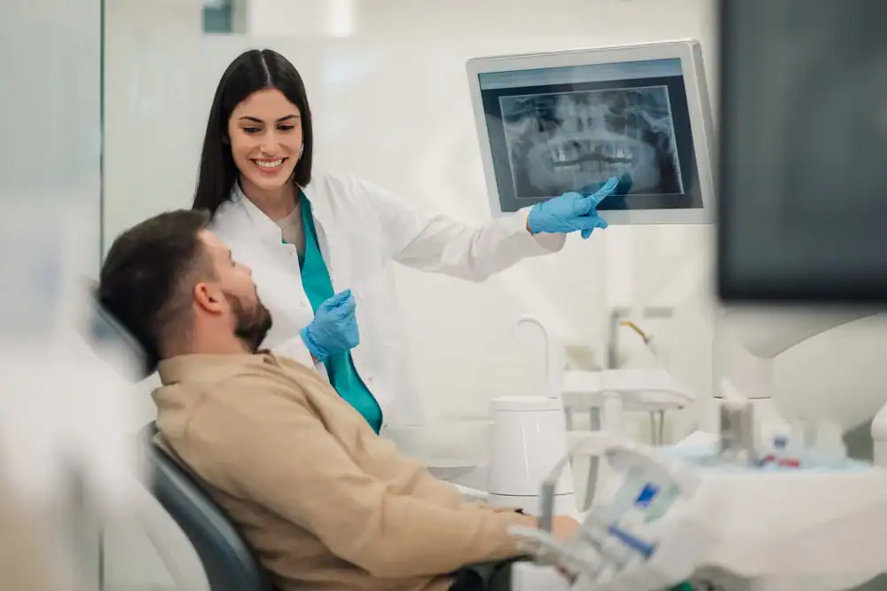 A dentist wearing gloves shows a dental X-ray to a male patient sitting in a dental chair. Both are smiling as the dentist points at the X-ray displayed on a monitor in a modern dental office.