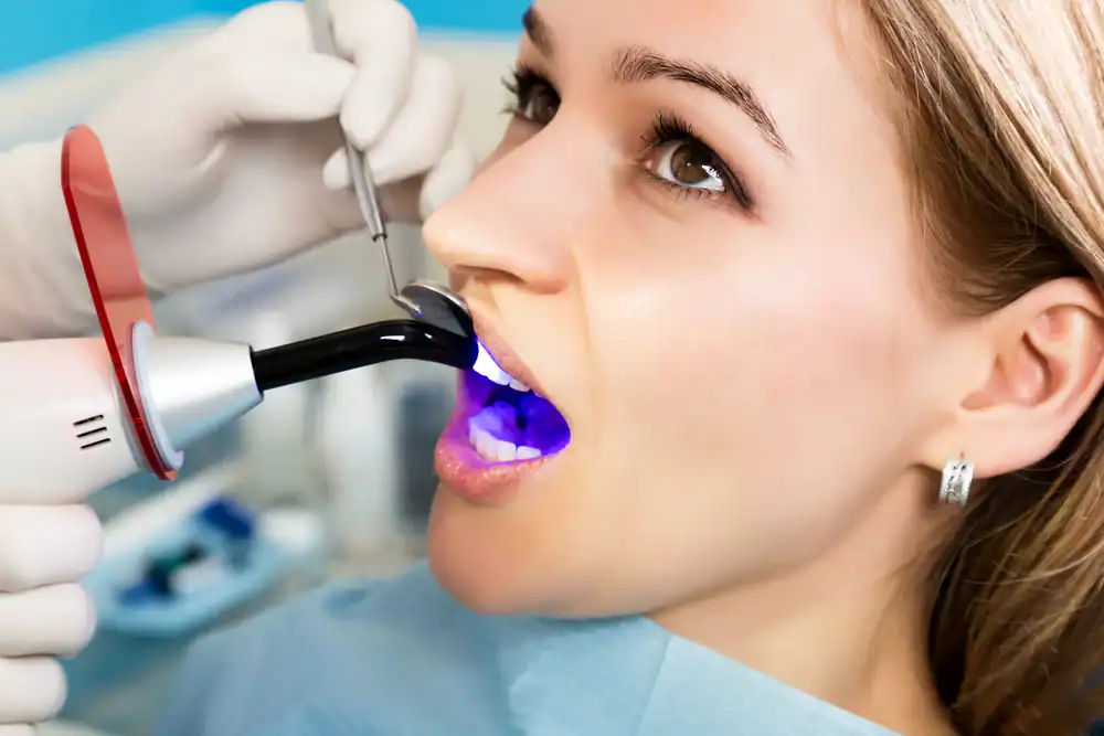 A woman sits in a dental chair with her mouth open as a dentist uses a curing light tool to harden dental material; the blue light shines inside her mouth while the dentist works carefully.