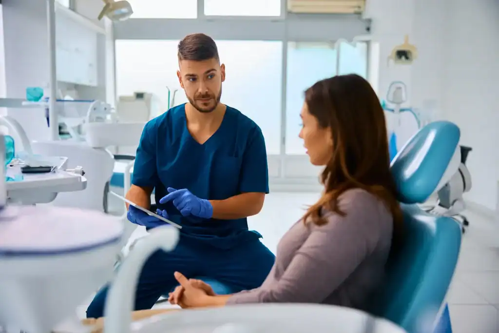 A dentist in blue scrubs and gloves discusses something with a female patient sitting in a dental chair inside a modern dentist clinic.