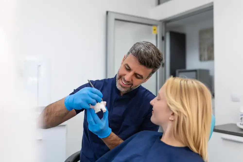 A dentist wearing blue gloves shows a dental model to a female patient in a dental clinic, explaining a procedure with a friendly smile, ensuring the patient feels comfortable and informed.