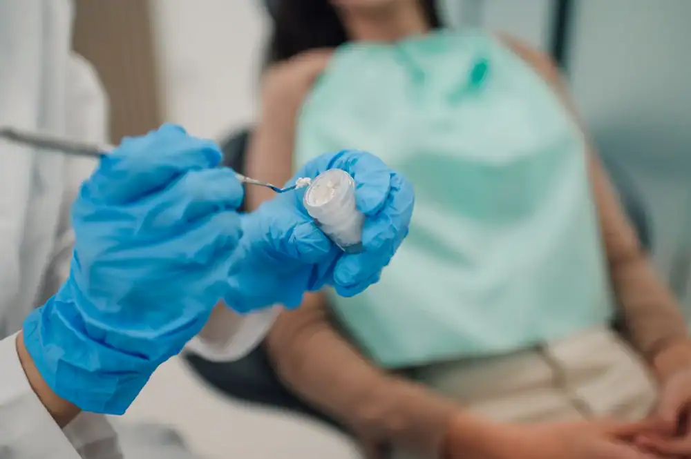 A dentist wearing blue gloves prepares dental material with a tool while a patient reclines in a dental chair, covered with a protective bib, as the dentist ensures comfort and precision.