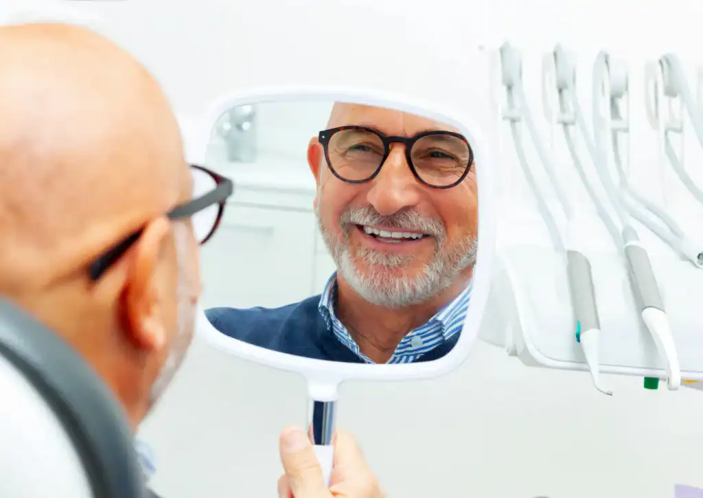 A smiling older man with glasses looks at his reflection in a handheld mirror while sitting in a dentist's office, surrounded by dental tools.
