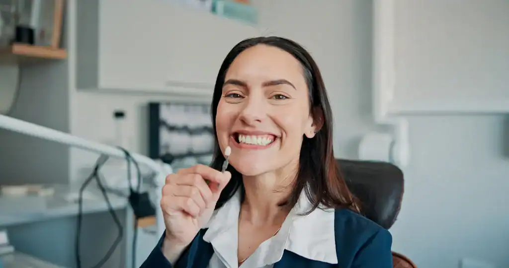 A woman at the dentist’s office smiles while holding a tooth shade guide to her teeth, likely for a color match or whitening consultation. She has dark hair and is wearing a blazer over a white shirt.