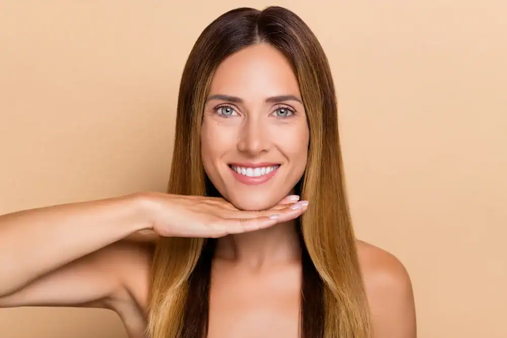 A smiling woman with straight, long brown hair holds her hand horizontally under her chin against a light beige background, showcasing a bright, dentist-approved smile.